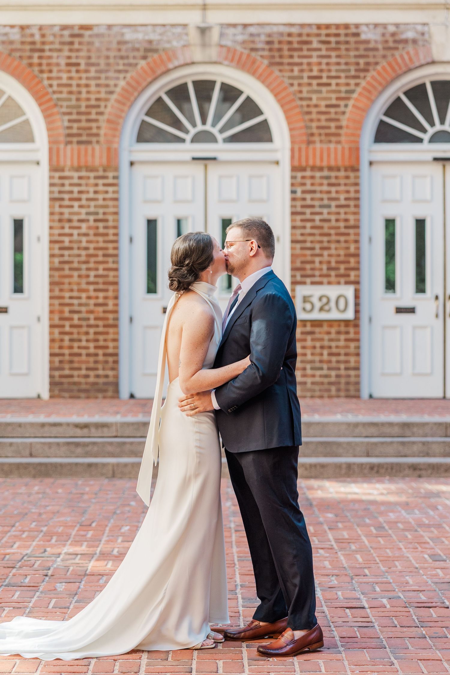 Bride and groom share a kiss in front of a brick building numbered 520, bride in elegant backless gown.