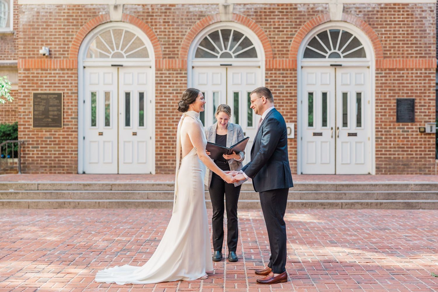 Couple exchanges vows with officiant outside brick building with white doors during outdoor wedding ceremony.