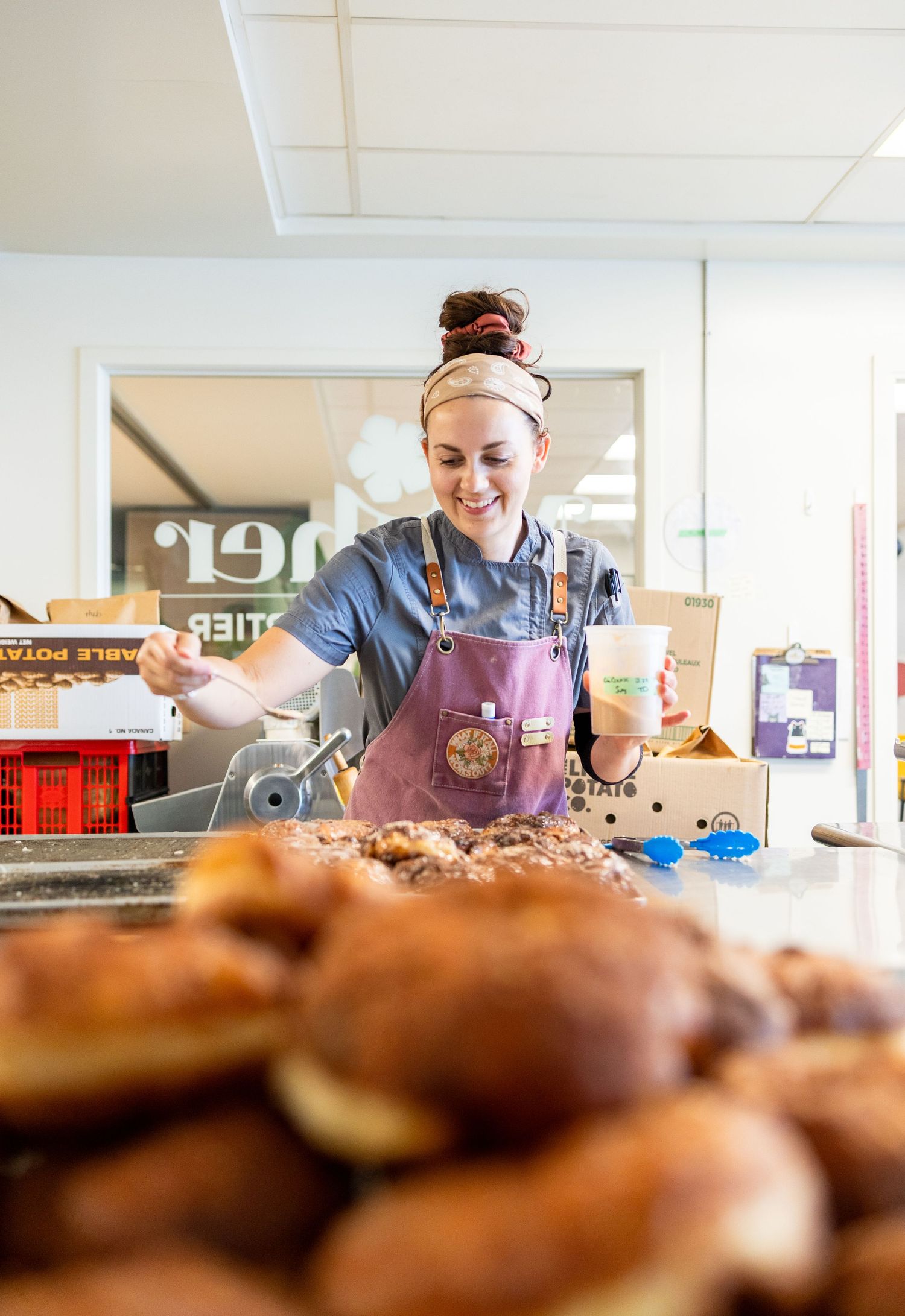 Smiling baker in apron arranges fresh pastries and donuts at a bakery counter while holding a coffee cup.