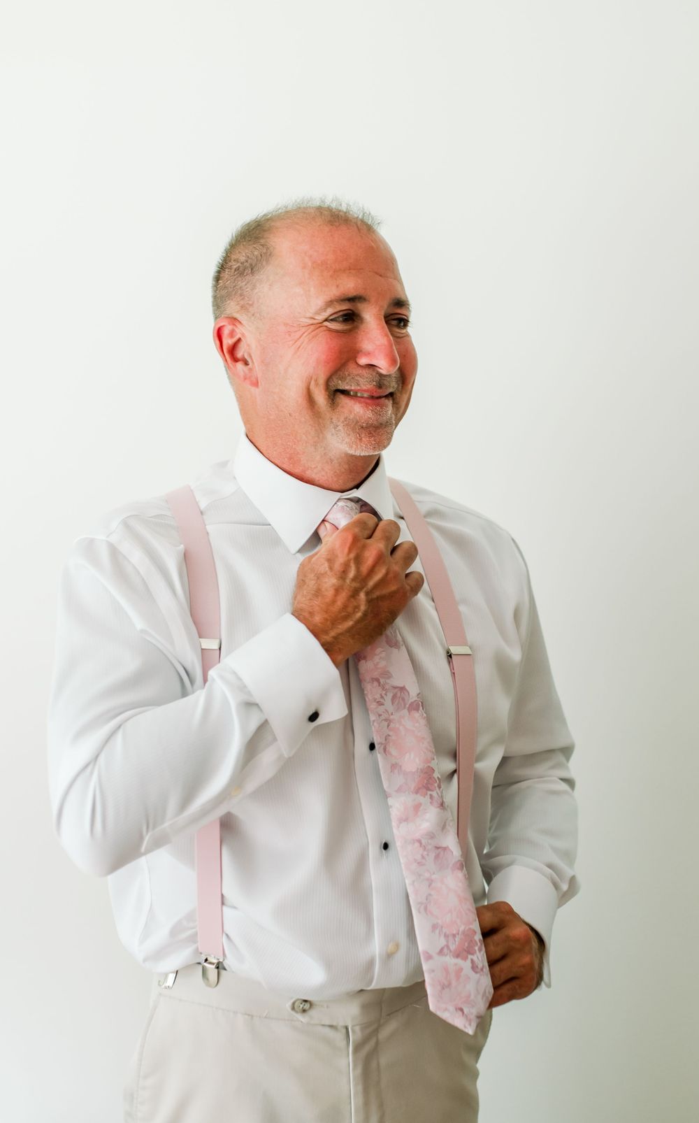 Smiling middle-aged man adjusting pink floral tie wearing white dress shirt with pink suspenders and khaki pants.