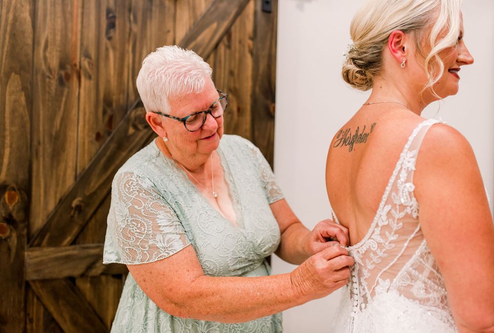 Older woman helping bride button lace wedding dress in rustic barn setting.