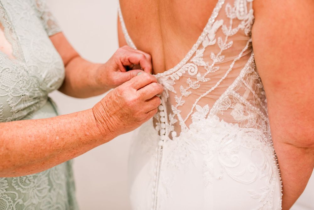 Bride having lace wedding dress buttoned up from behind by an older woman helping with bridal preparations.