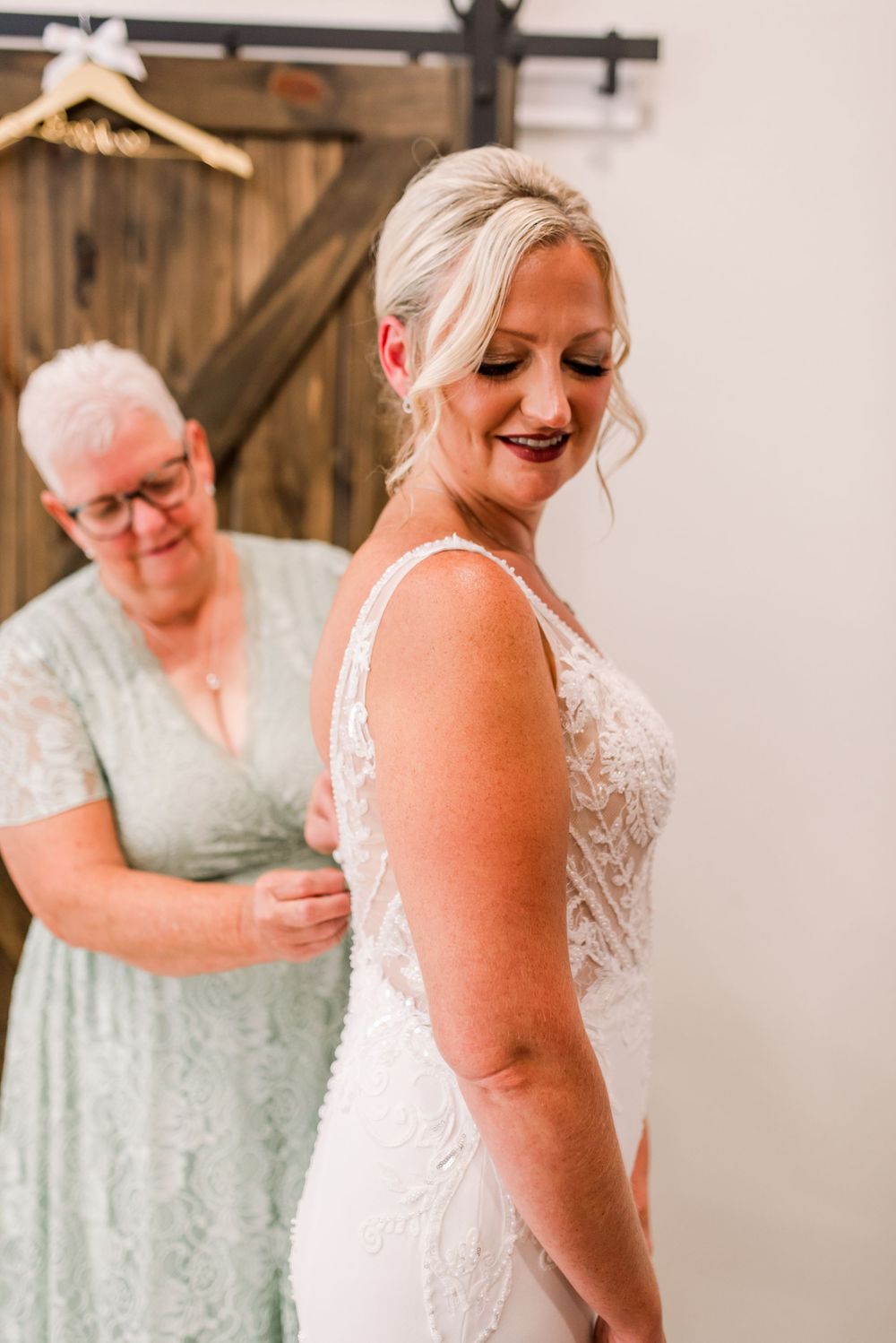 Bride in lace wedding gown smiling as older woman in sage green dress fastens the back near rustic barn doors.