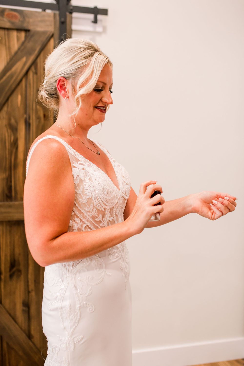 Bride in lace wedding gown applying perfume near rustic barn doors before her ceremony.