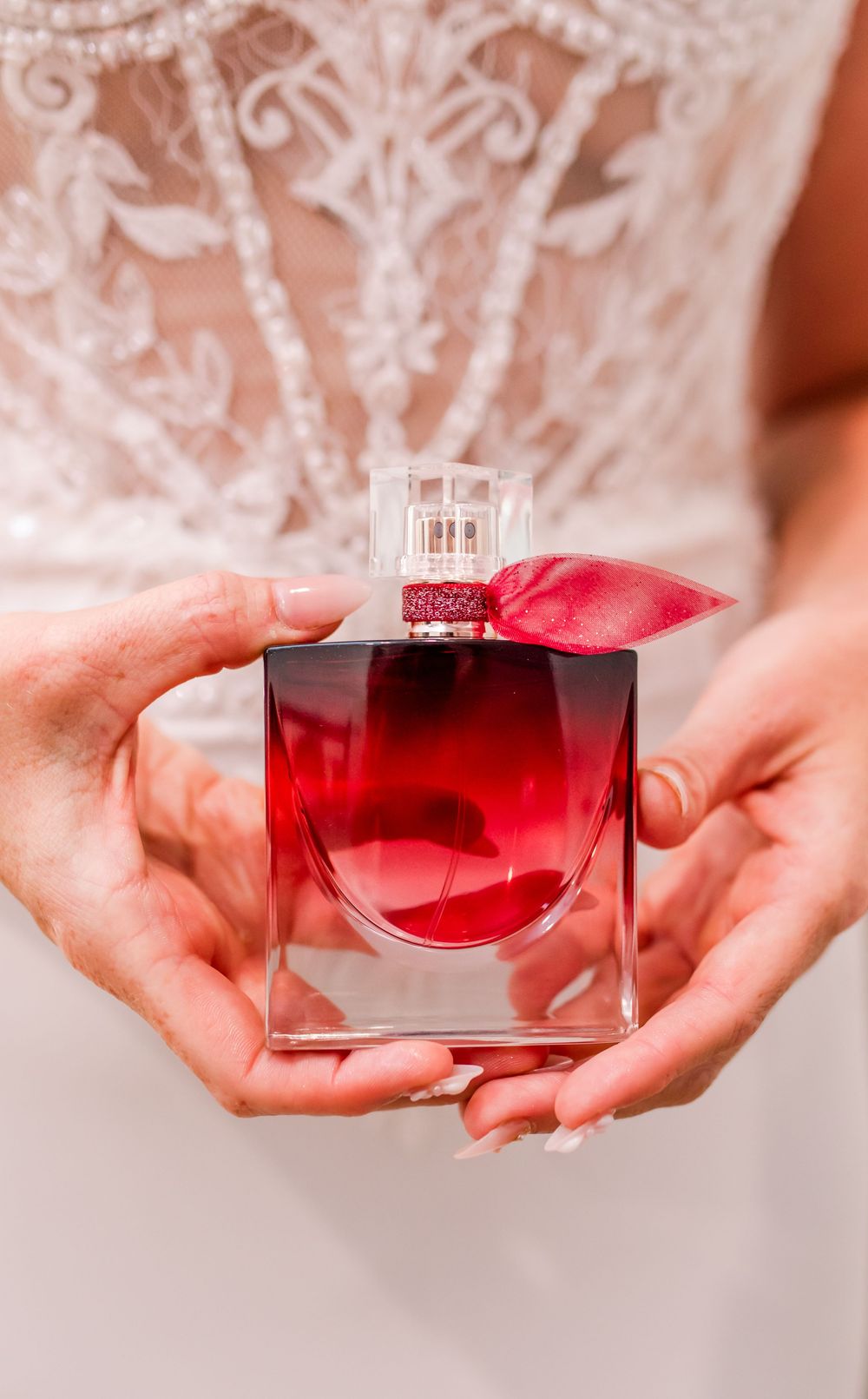 Bride holds a red Lancome perfume bottle with pink bow against her beaded white wedding gown.