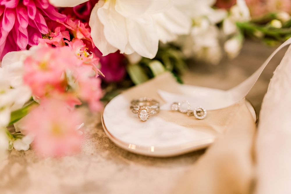 Close-up of a diamond engagement ring and wedding band resting on a bridal shoe near pink flowers.