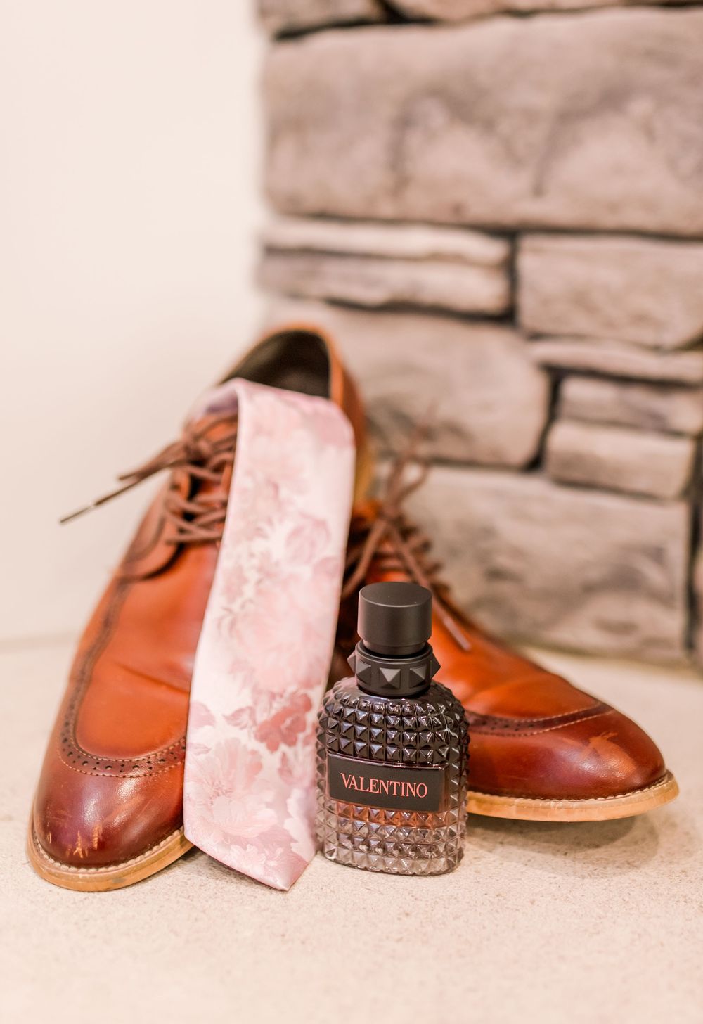Brown leather dress shoes with a floral pink tie and Valentino cologne bottle arranged for a wedding flat lay.