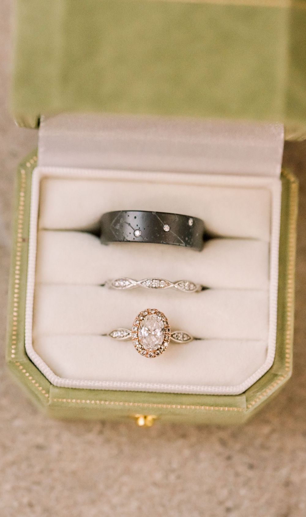 Three wedding rings displayed in a vintage green ring box, featuring a dark mens band, diamond eternity band, and oval halo engagement ring.
