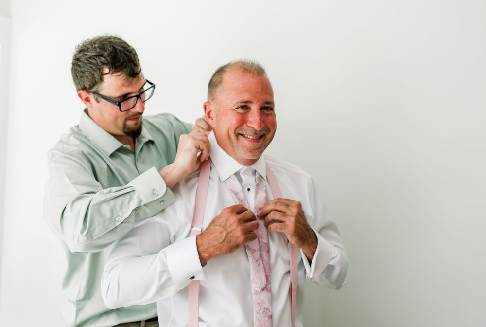 Man helping smiling groom adjust pink tie and shirt collar before wedding ceremony indoors.