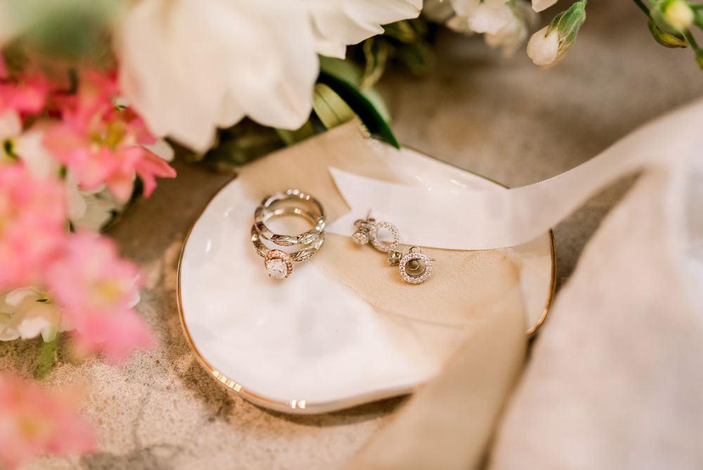 Gold wedding rings and pearl drop earrings displayed on a white dish surrounded by pink and white wedding flowers.