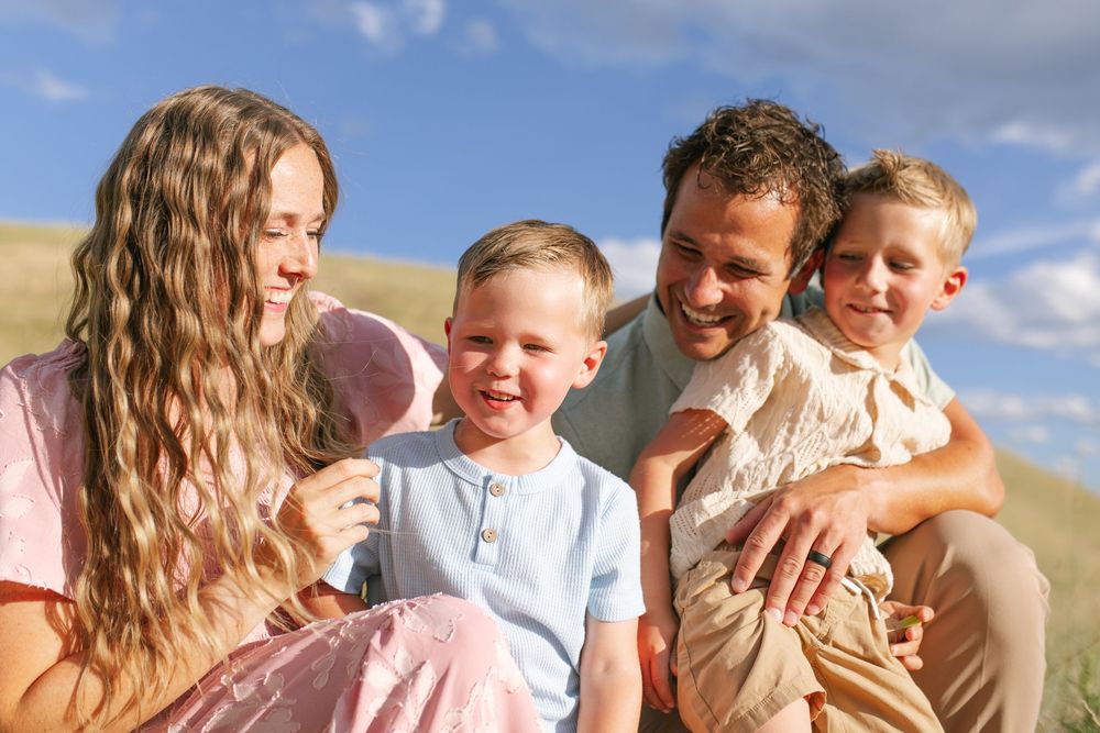 Smiling family of four sitting together outdoors in sunny grassy hills, sharing a joyful moment.
