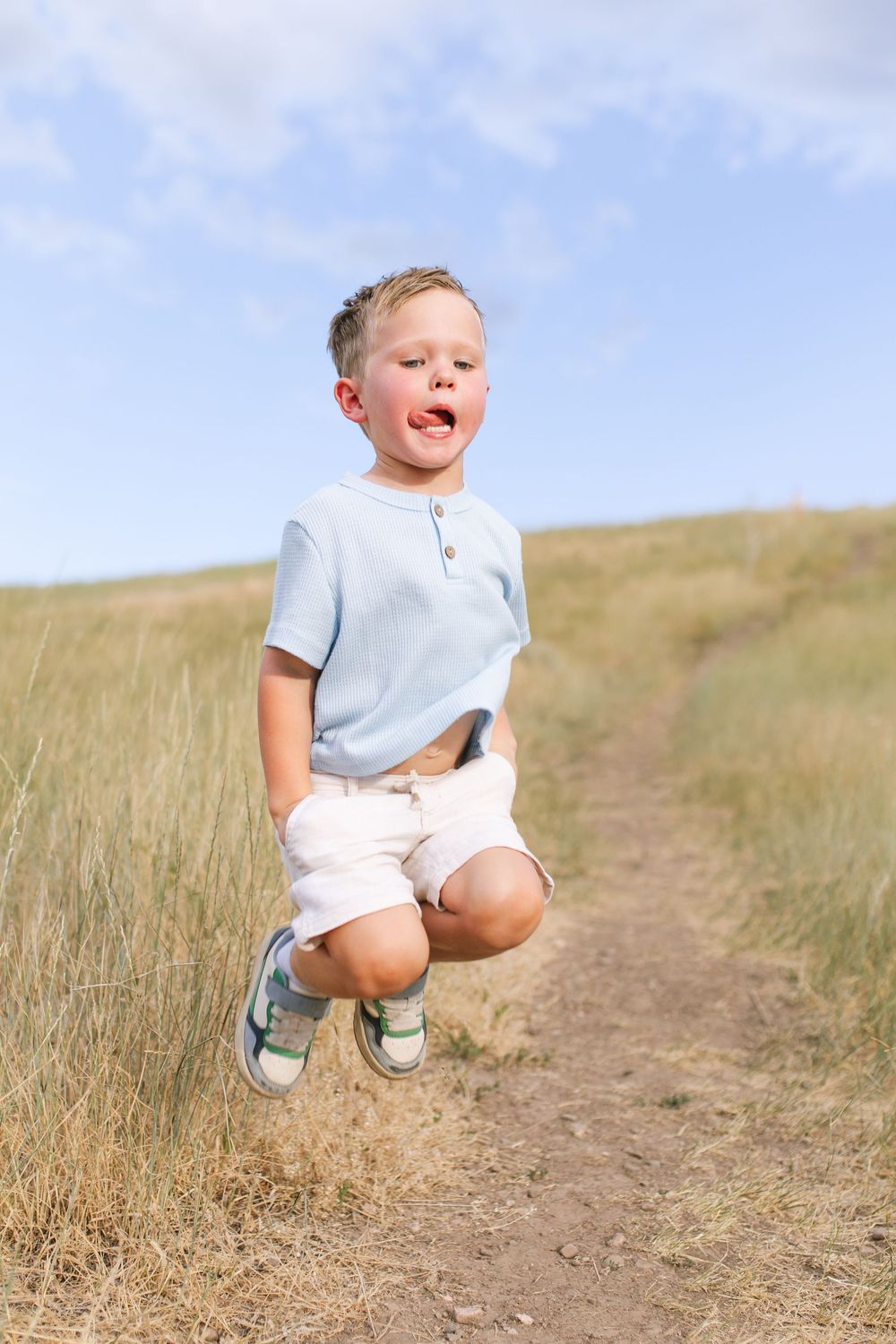 A young boy jumps with excitement on a dirt path through golden grass fields under a blue sky.