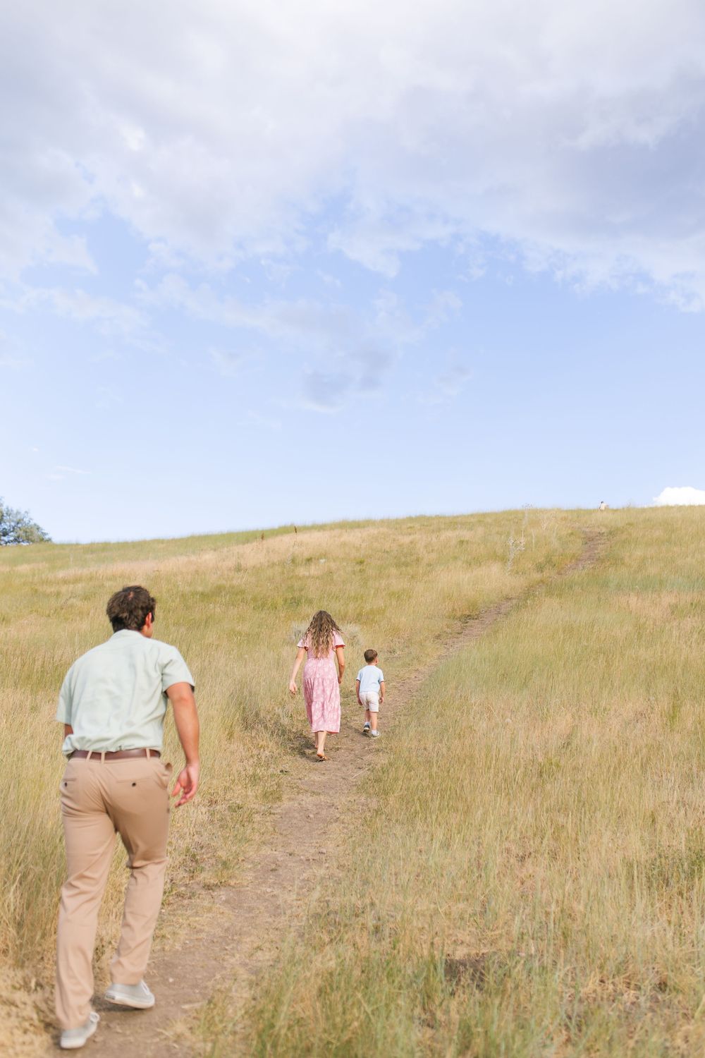 A father walks with two young children along a grassy trail under a partly cloudy sky.