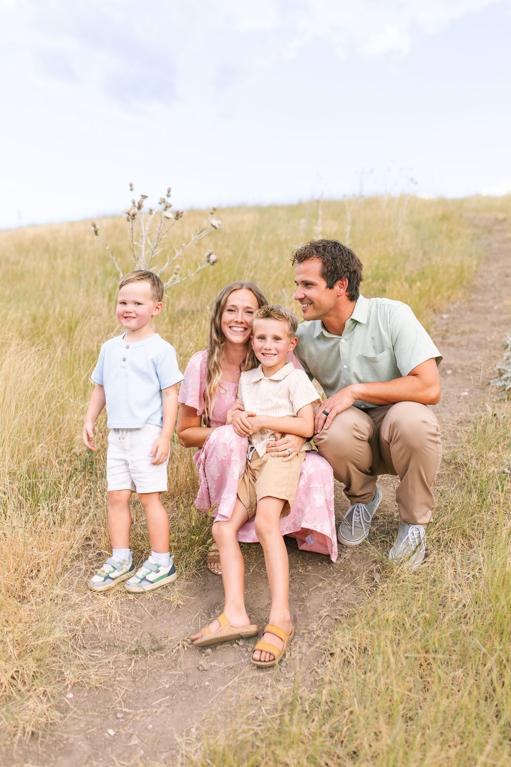 Happy family of four poses on a scenic dirt path surrounded by golden grass fields.