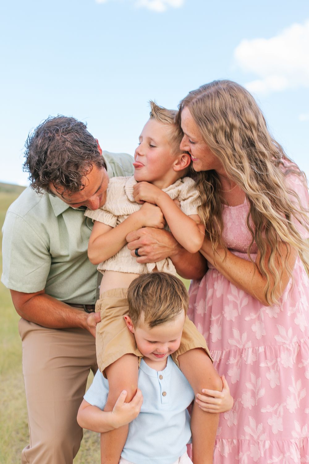 Happy family of four sharing hugs and kisses outdoors in a grassy field under blue sky.