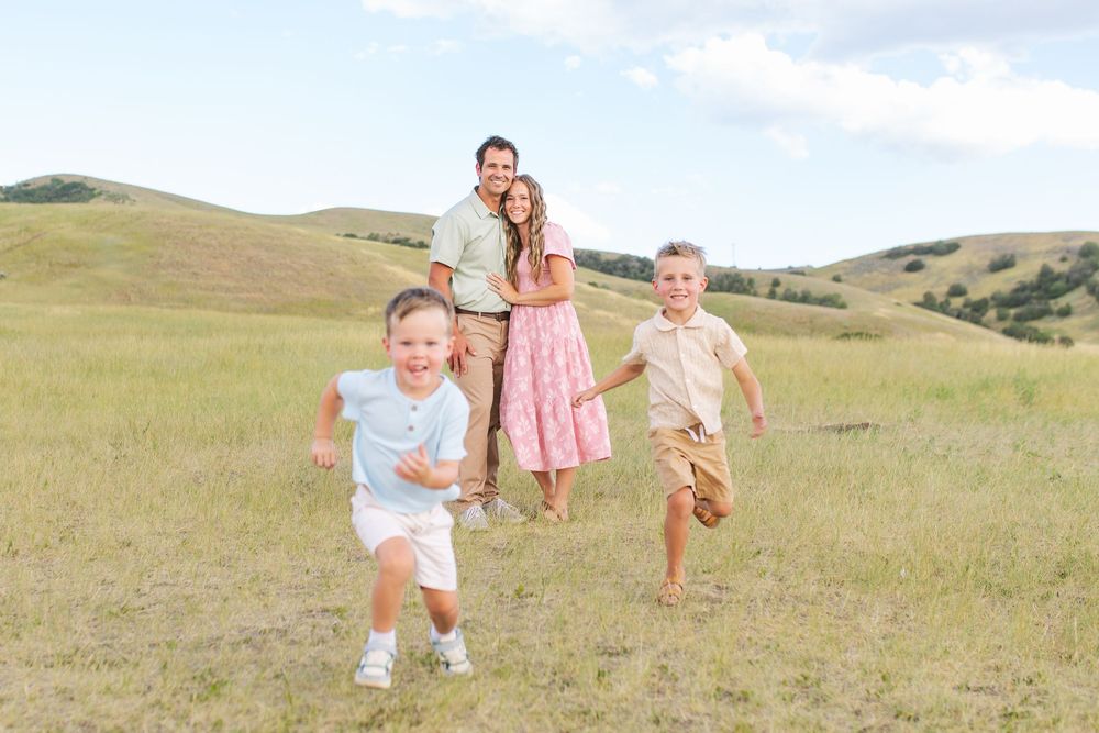 Joyful family with two young boys running toward camera in a scenic green hillside meadow.