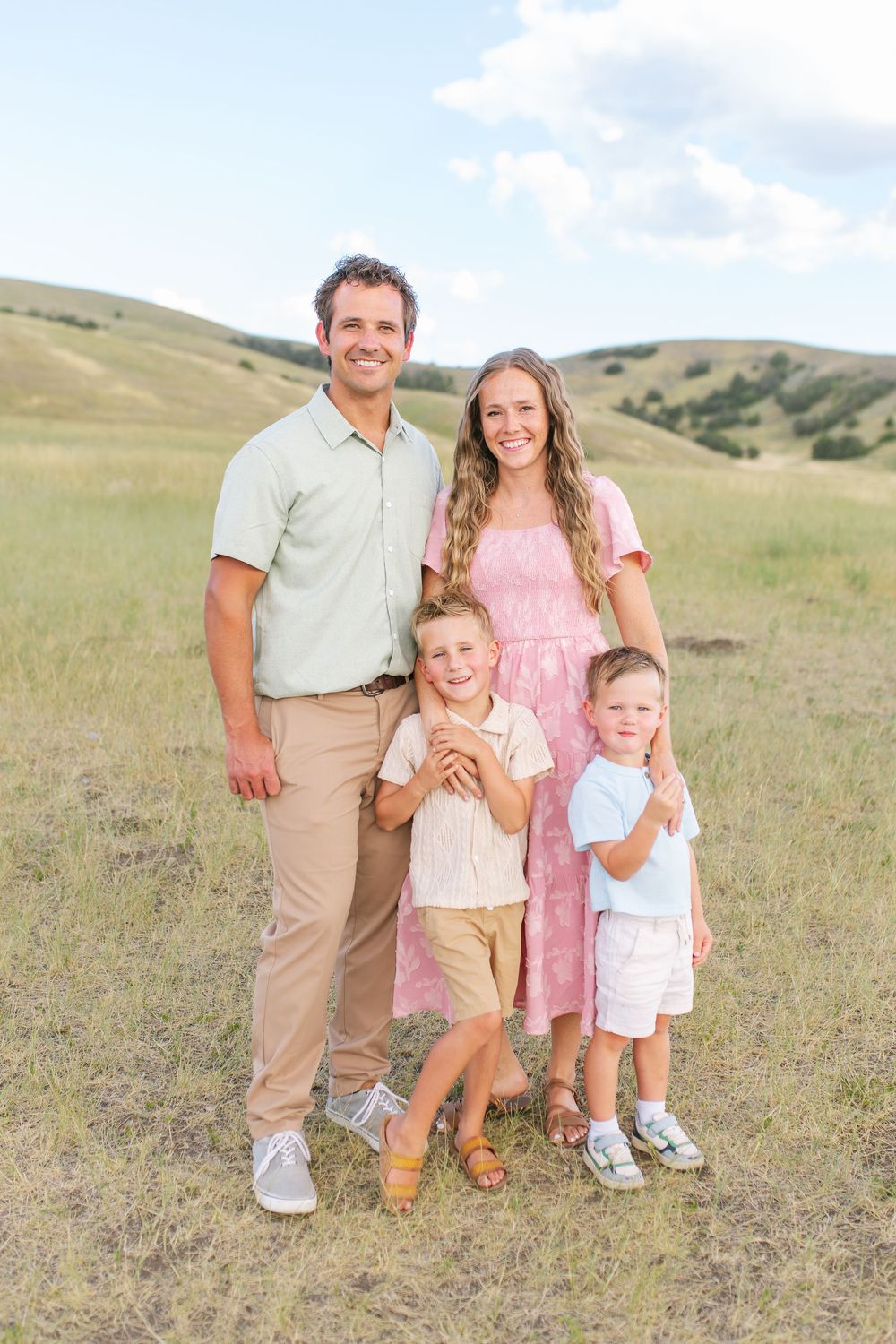A smiling family of four poses in a green meadow, parents with two young boys in coordinated outfits.