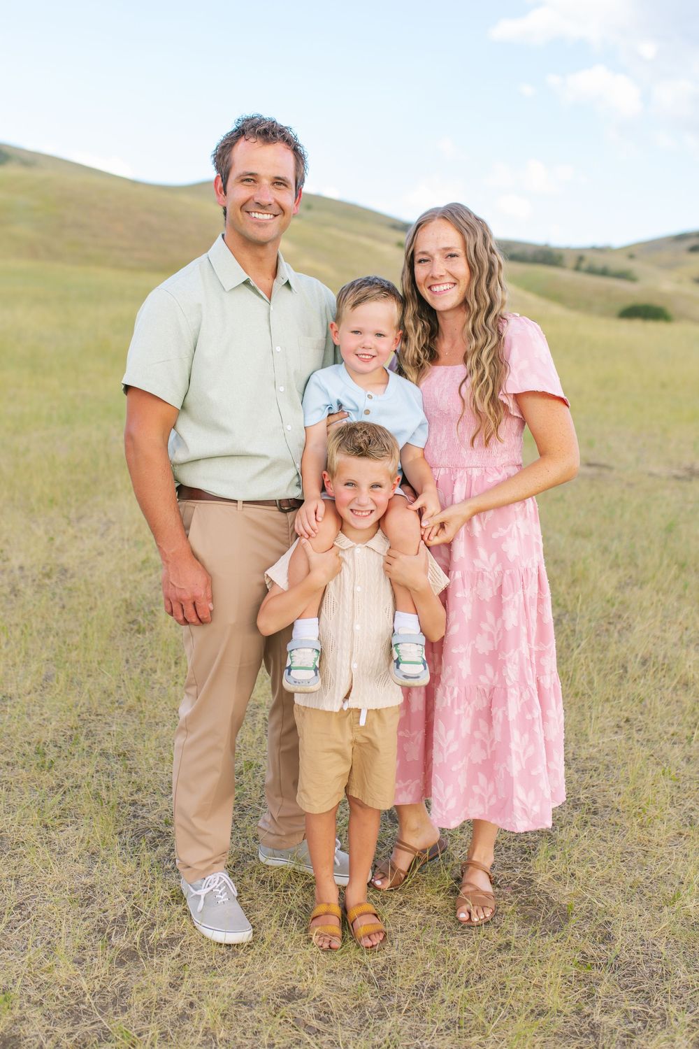 Happy family of four posing outdoors in a green meadow, parents with two young boys in coordinated pastel outfits.