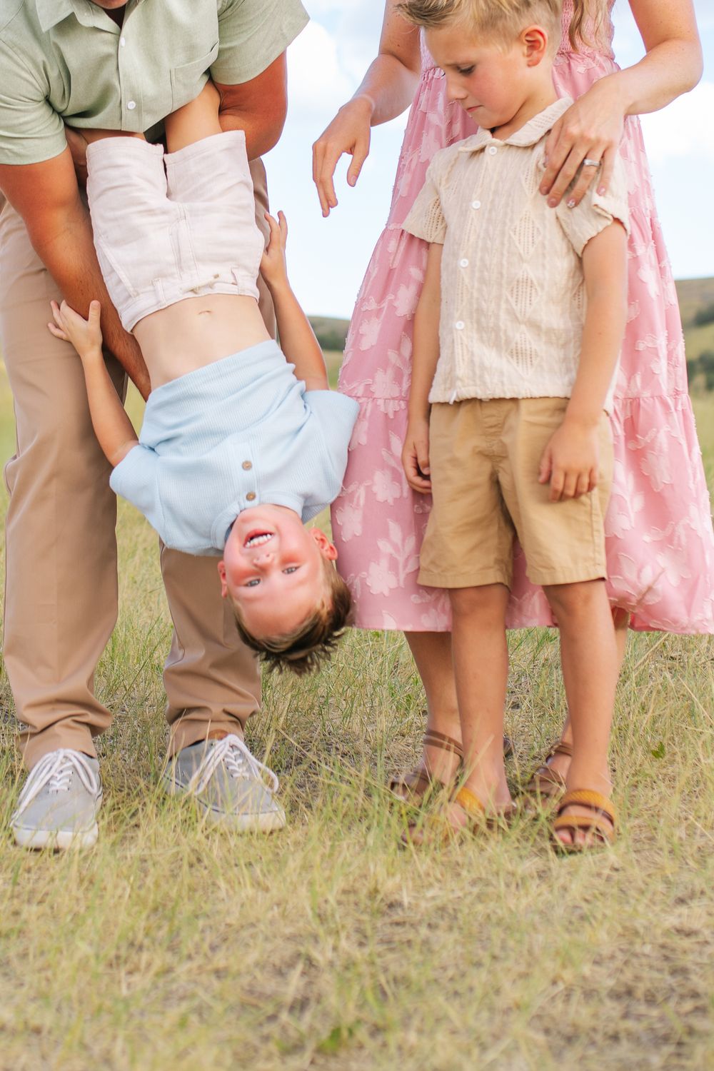 Father playfully holds young boy upside down while mother steadies older child in grassy field.