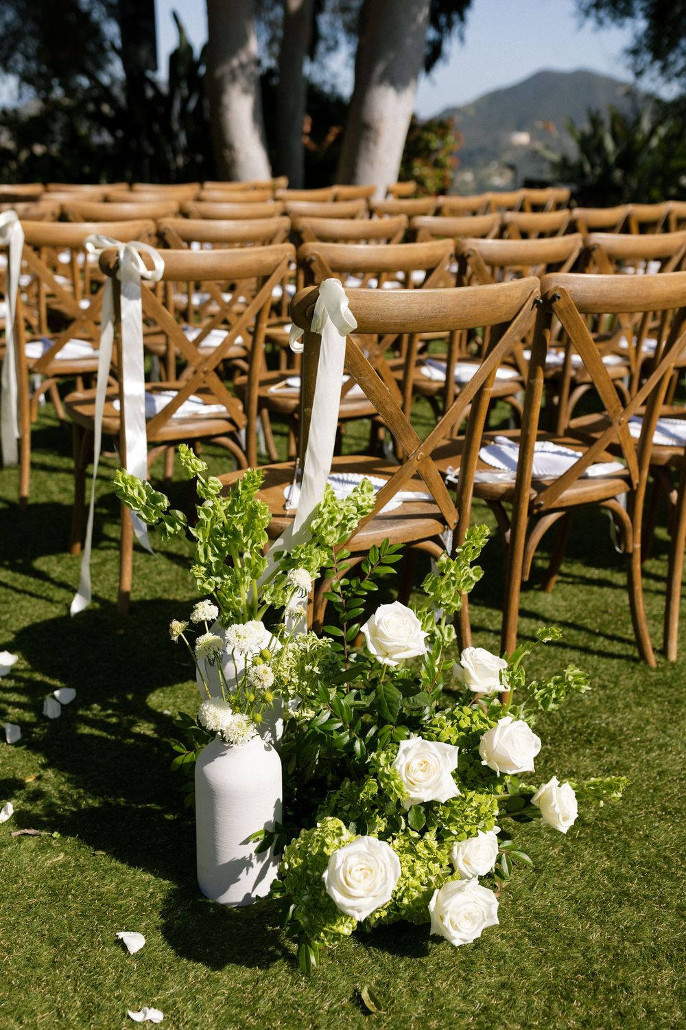 White roses and greenery floral arrangement beside wooden cross-back chairs at an outdoor wedding ceremony.