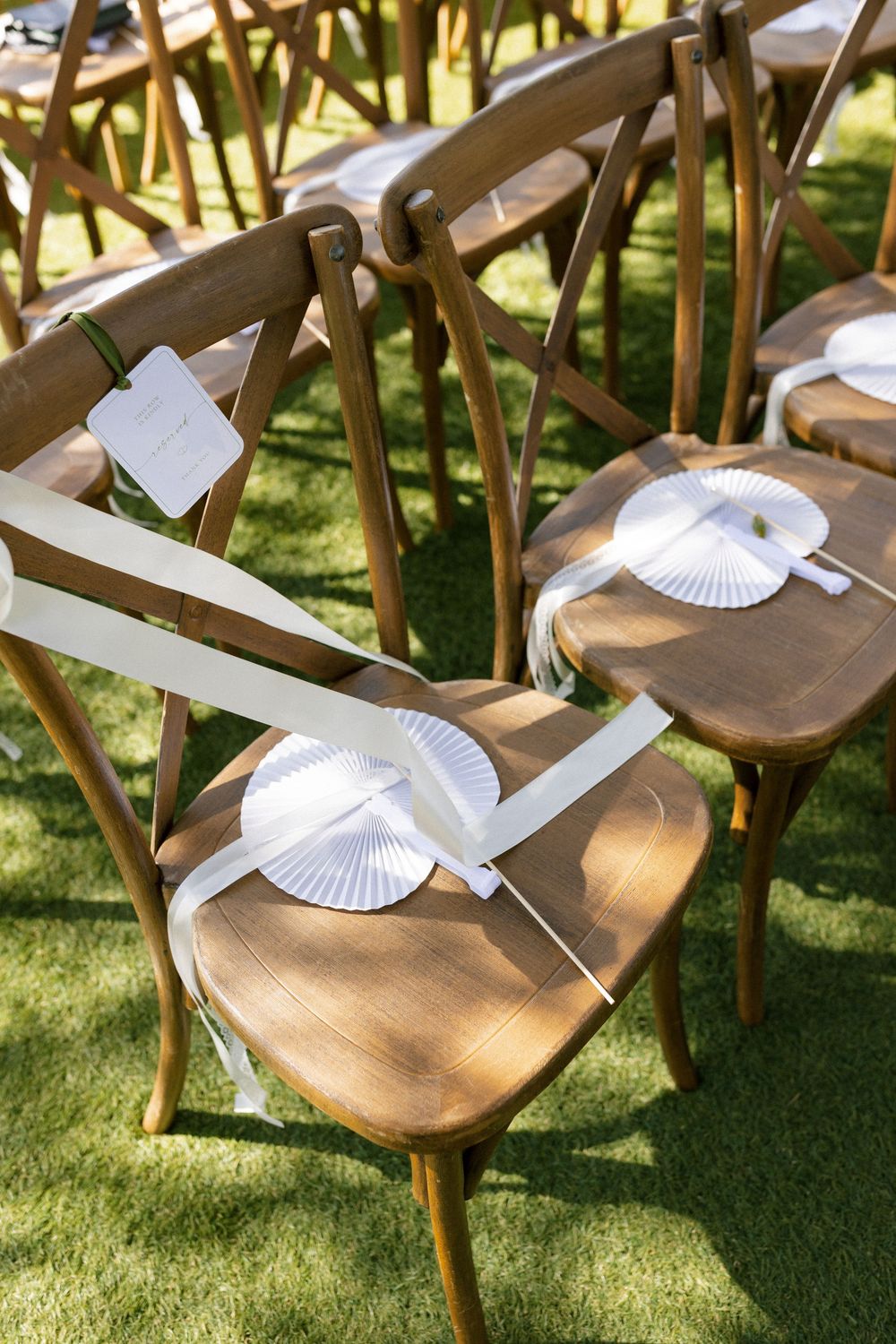 Wooden cross-back chairs with white paper fans and ribbon tied to seats arranged on green grass for an outdoor wedding ceremony.