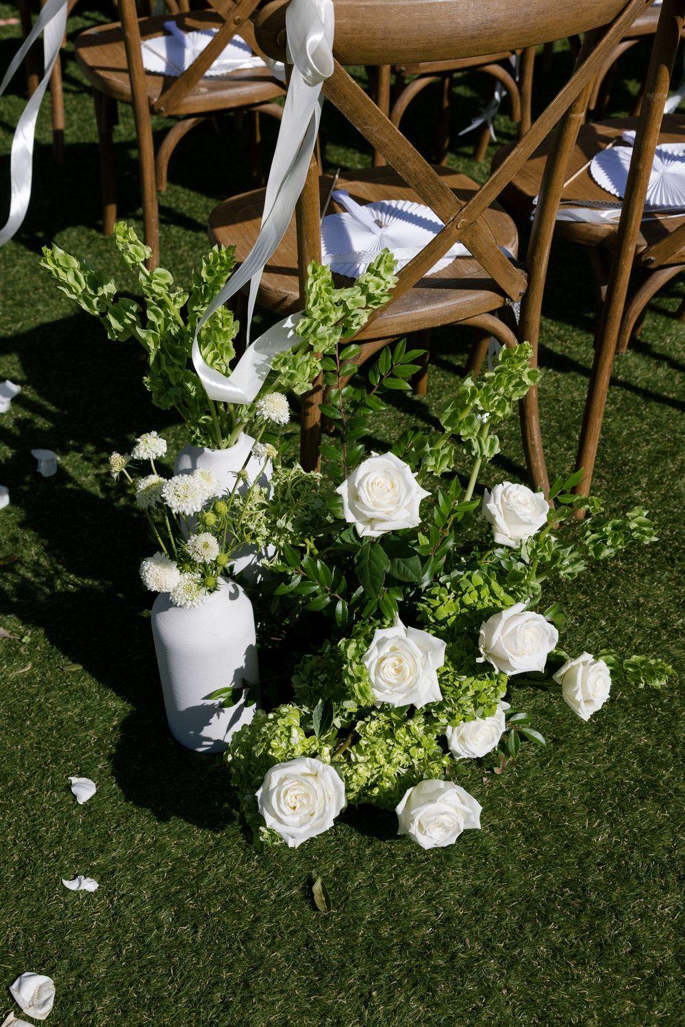 White roses and green hydrangea floral arrangements beside wooden cross-back chairs at outdoor wedding.