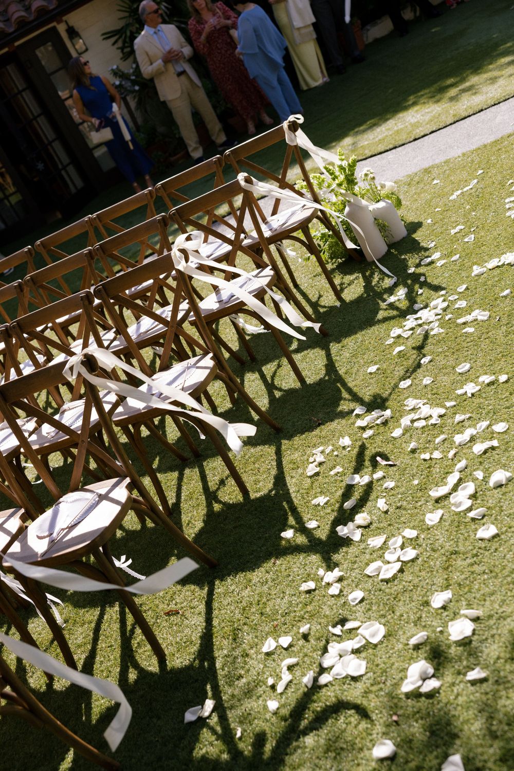 Wooden chairs with white ribbon bows line a grass aisle scattered with flower petals at an outdoor wedding ceremony.