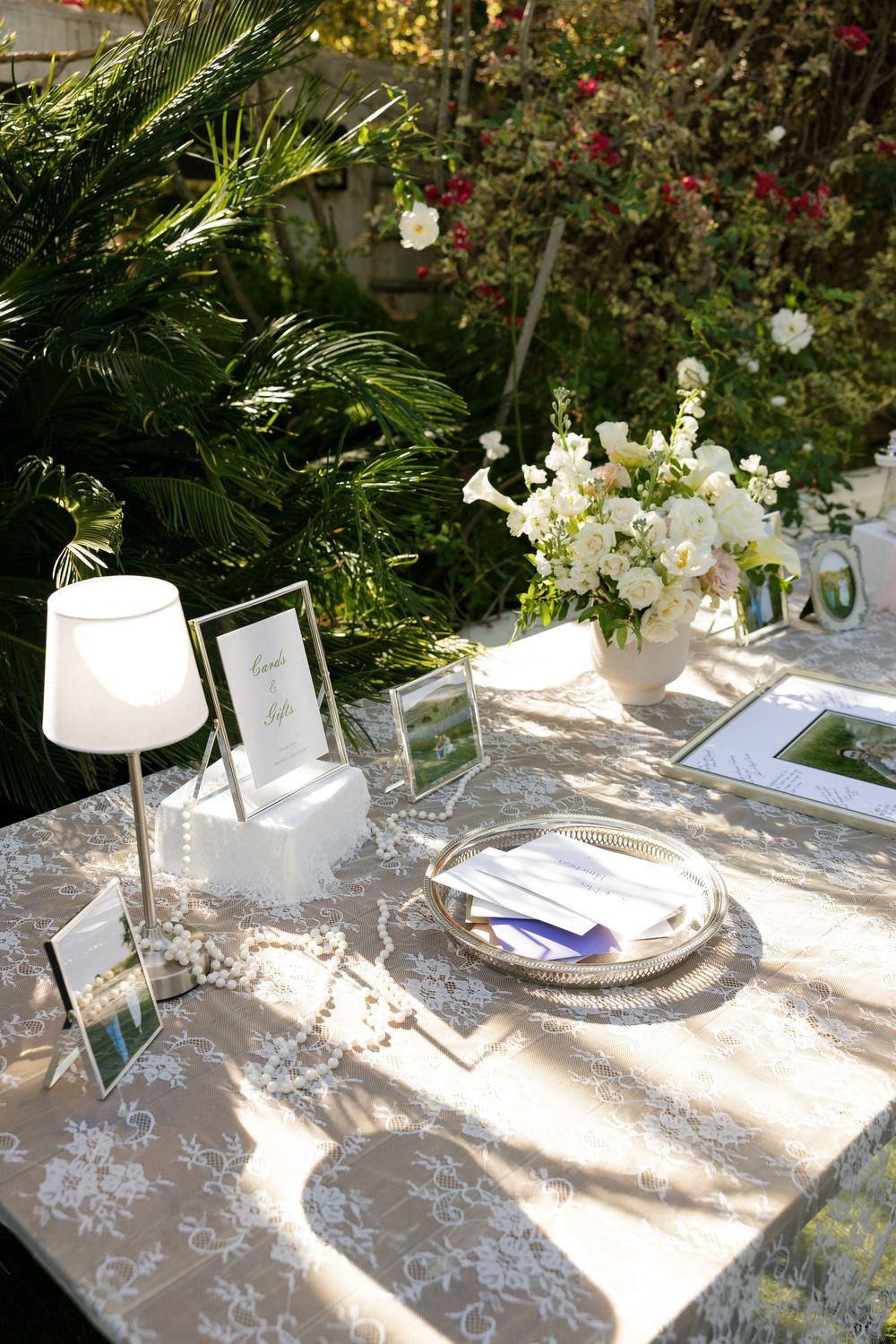Elegant outdoor wedding reception table with lace tablecloth, white floral centerpiece, silver tray, and framed signs in a garden setting.