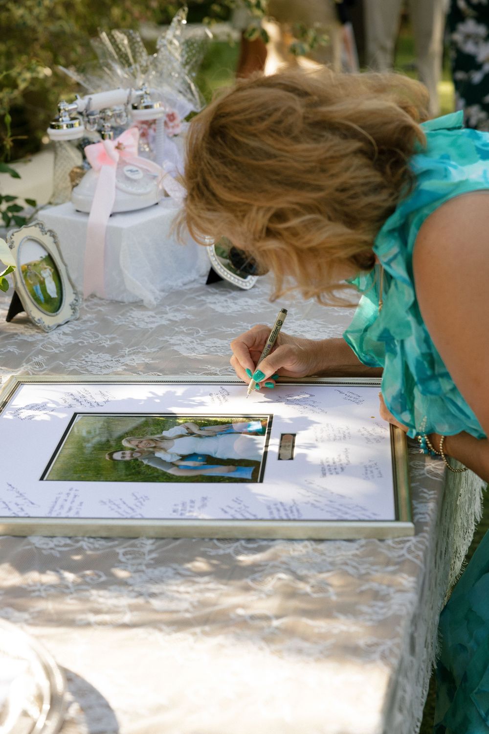 Woman in teal dress signing a framed wedding guest book with photo at an outdoor reception table.