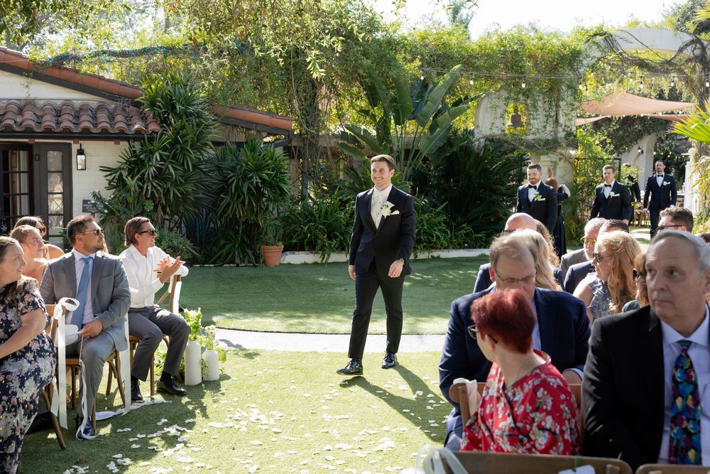 Groom standing at outdoor garden wedding ceremony aisle with seated guests on both sides.