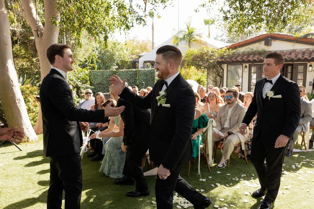 Groomsmen in black tuxedos greeting each other at an outdoor wedding ceremony with seated guests.
