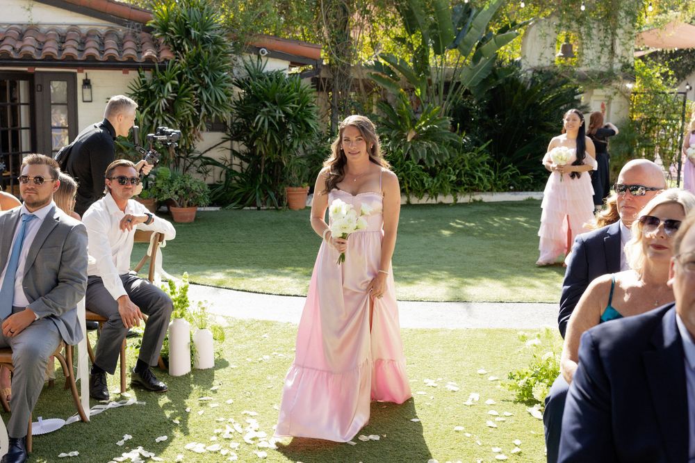 Bridesmaid in pink gown walking down outdoor wedding ceremony aisle carrying white bouquet.