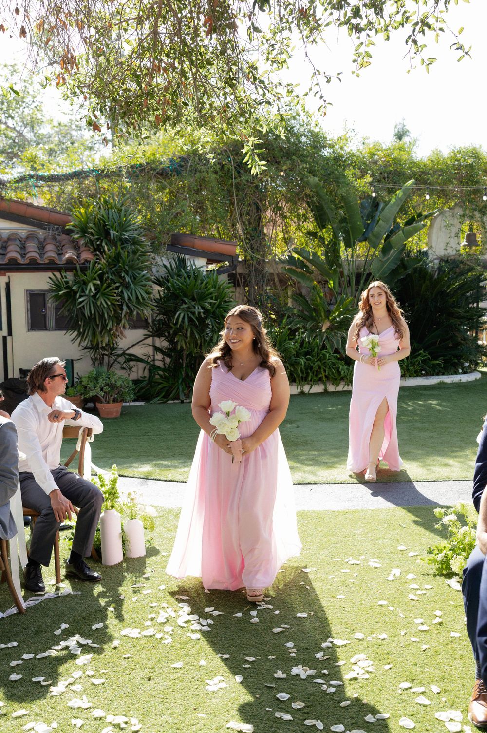 Bridesmaids in pink dresses walk down outdoor wedding aisle on green lawn with flower petals and lush garden backdrop.