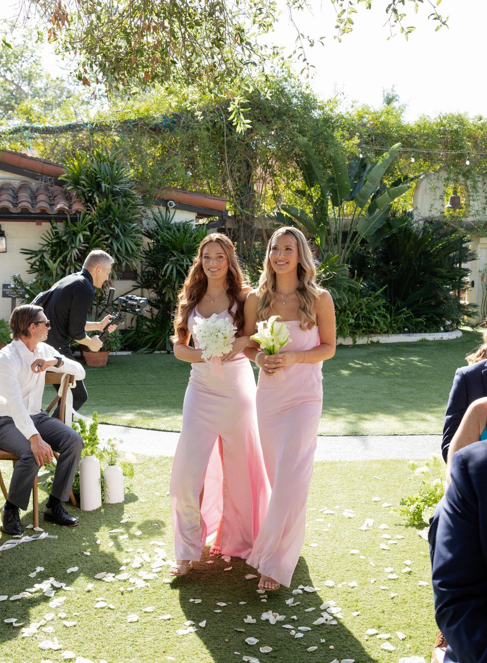 Two bridesmaids in pink gowns walk down outdoor wedding aisle carrying white bouquets.