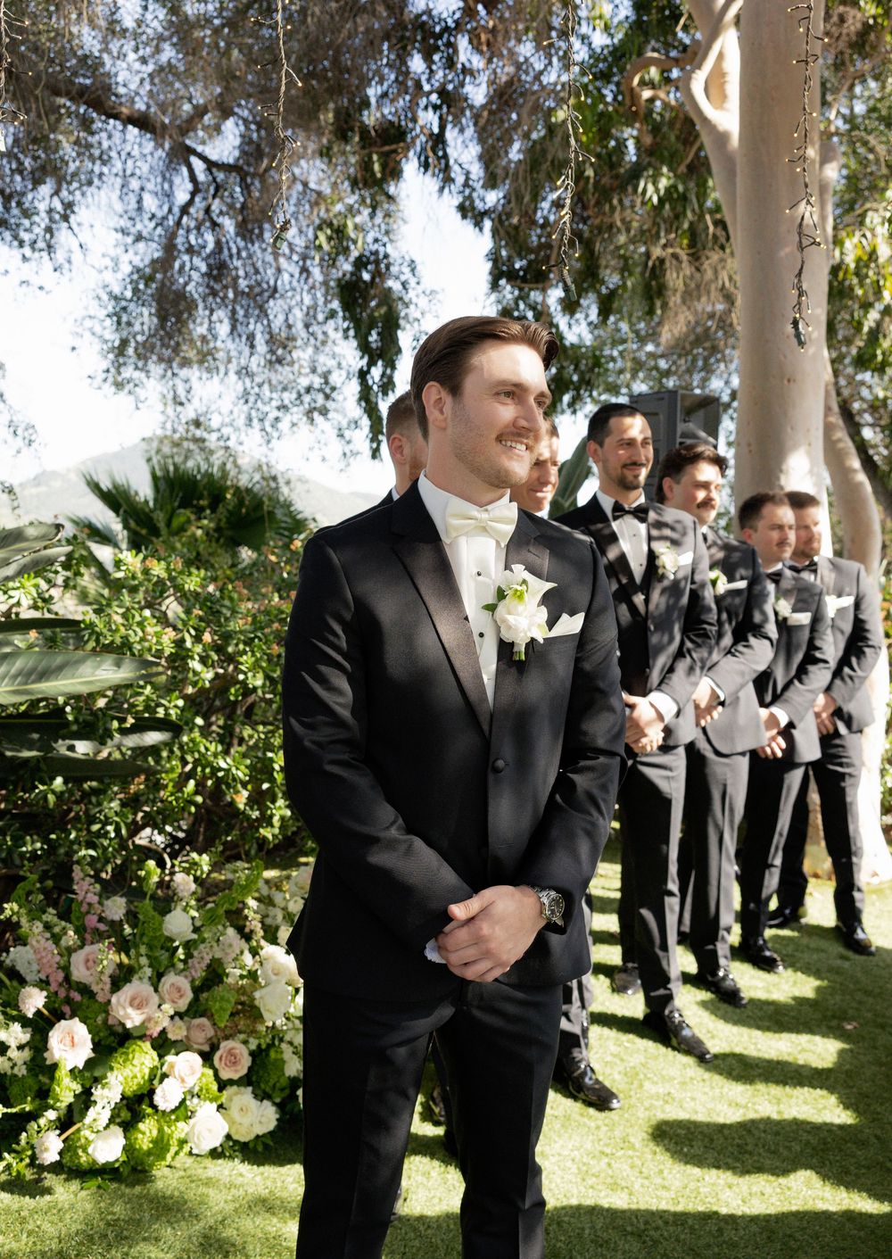 Groom in black tuxedo smiling at altar with groomsmen in gray suits lined up at outdoor wedding ceremony.