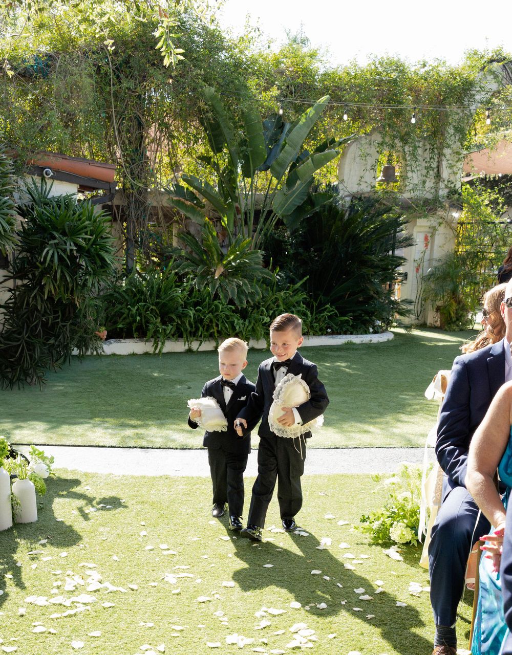 Two young ring bearers in tuxedos walk down the aisle at an outdoor wedding ceremony surrounded by lush greenery.