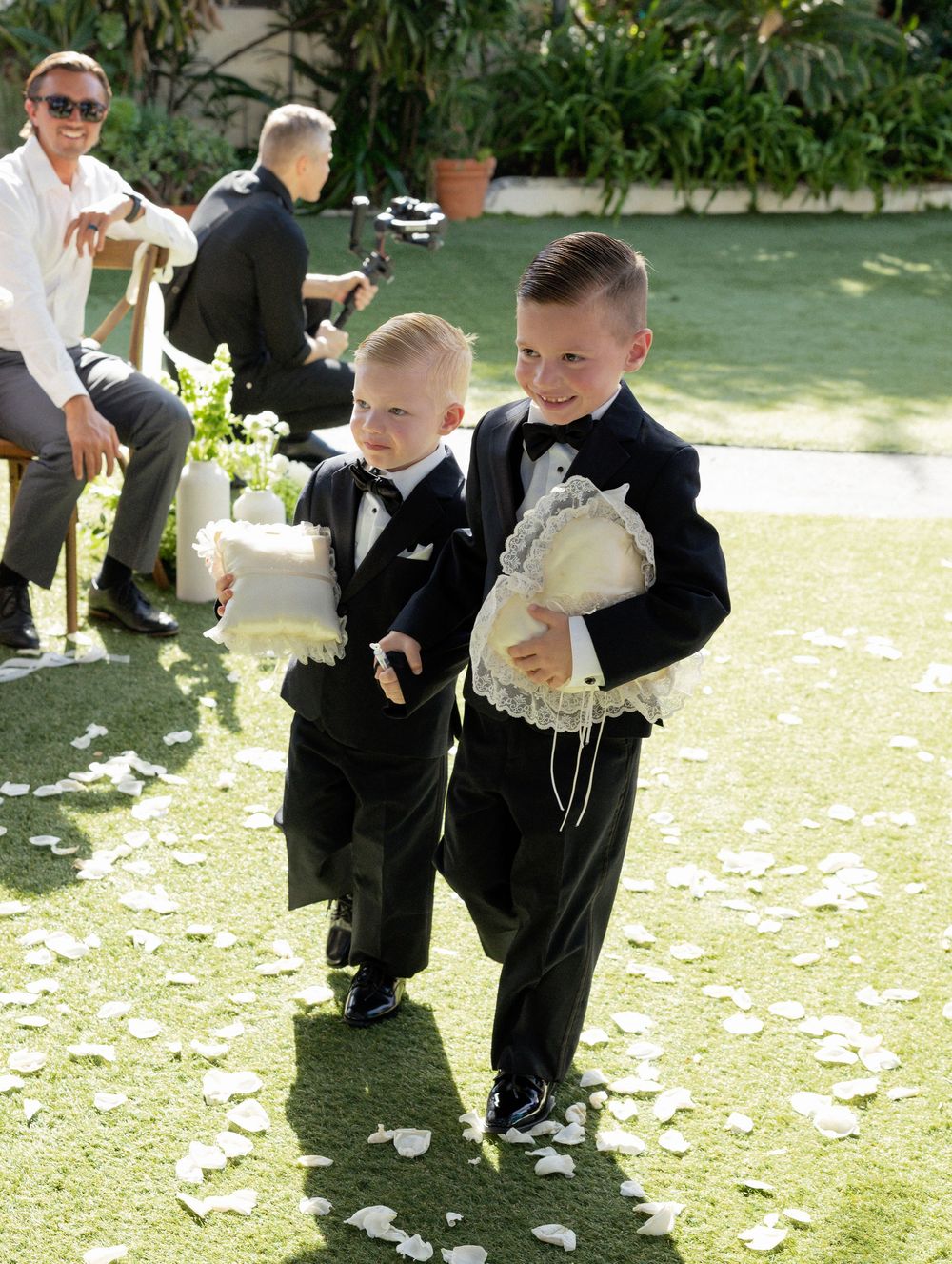 Two young ring bearers in black tuxedos carry white pillows down a flower petal-lined aisle at an outdoor wedding.