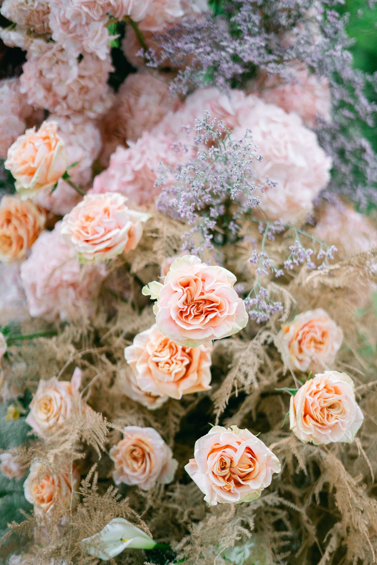 Lush arrangement of peach roses, pink peonies, and lavender wildflowers with dried grasses in a romantic floral display.