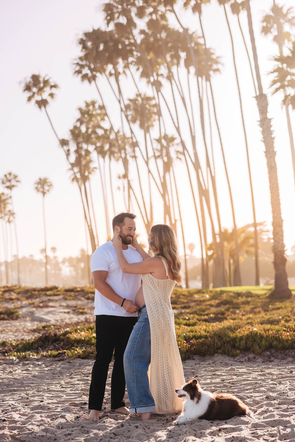 Couple embracing on sandy beach at golden hour with tall palm trees and a dog nearby.