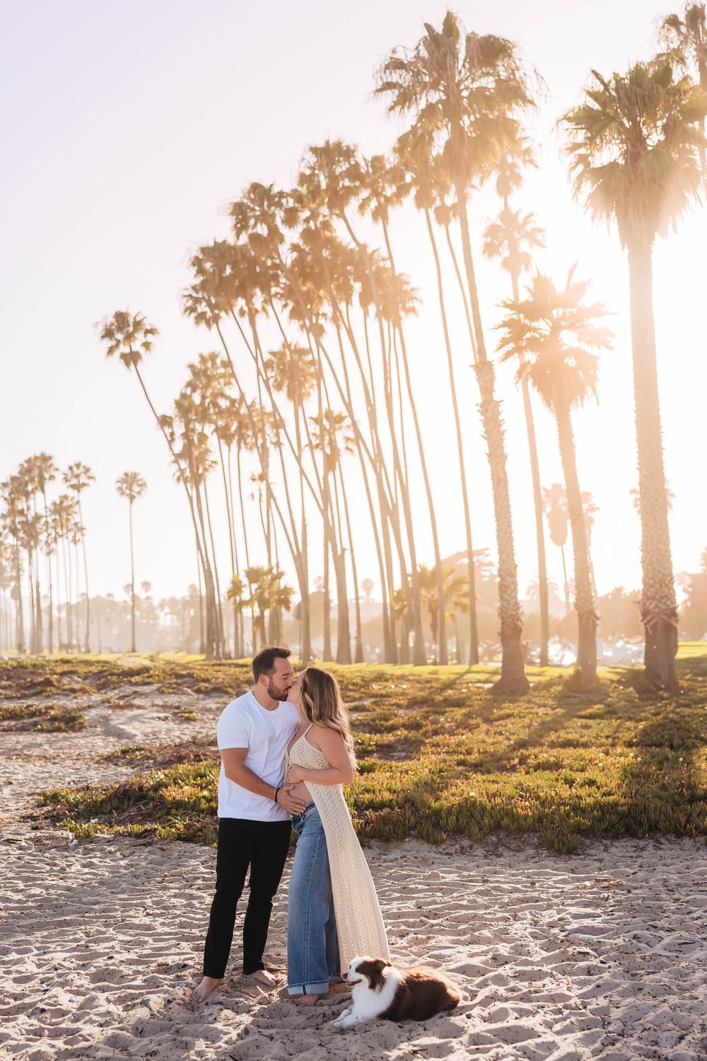 Couple embracing on sandy beach at golden hour with tall palm trees glowing in warm sunlight and a dog resting nearby.