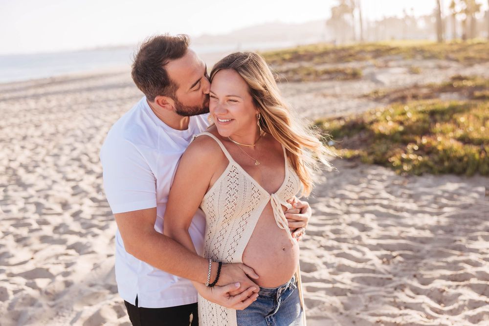 A couple embraces on a sunny beach, the woman visibly pregnant, as the man kisses her cheek tenderly. What to Wear
