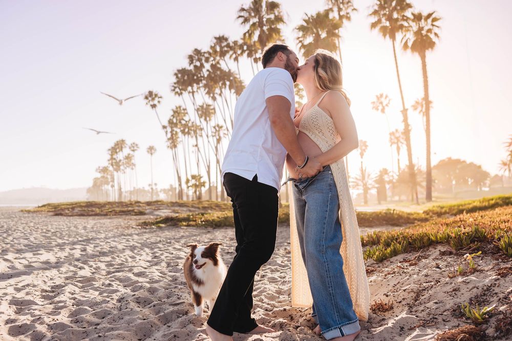 Couple kissing on a sunny beach with palm trees, jeans, white top, and a dog nearby. Santa Barbara maternity photos