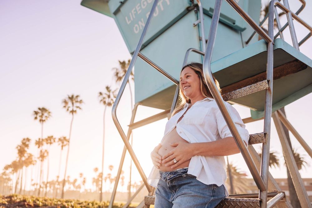 Pregnant woman in white top and jeans leaning against a lifeguard tower at sunset with palm trees in background.
