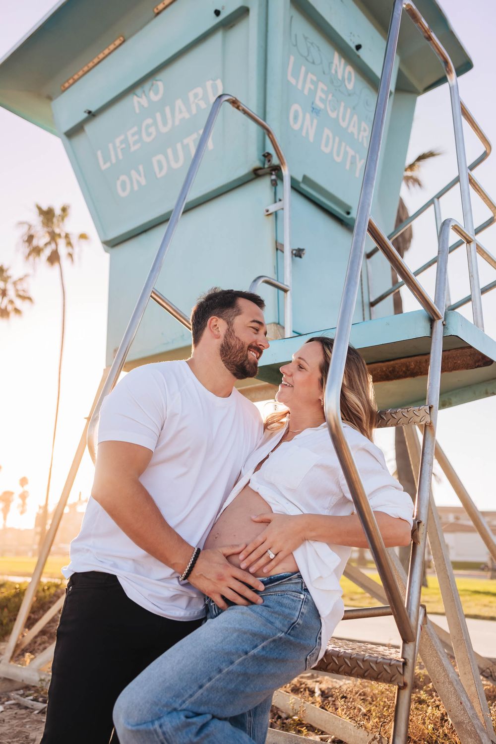 Happy couple in matching white tops cradle pregnant belly by a teal lifeguard tower at golden hour beach.