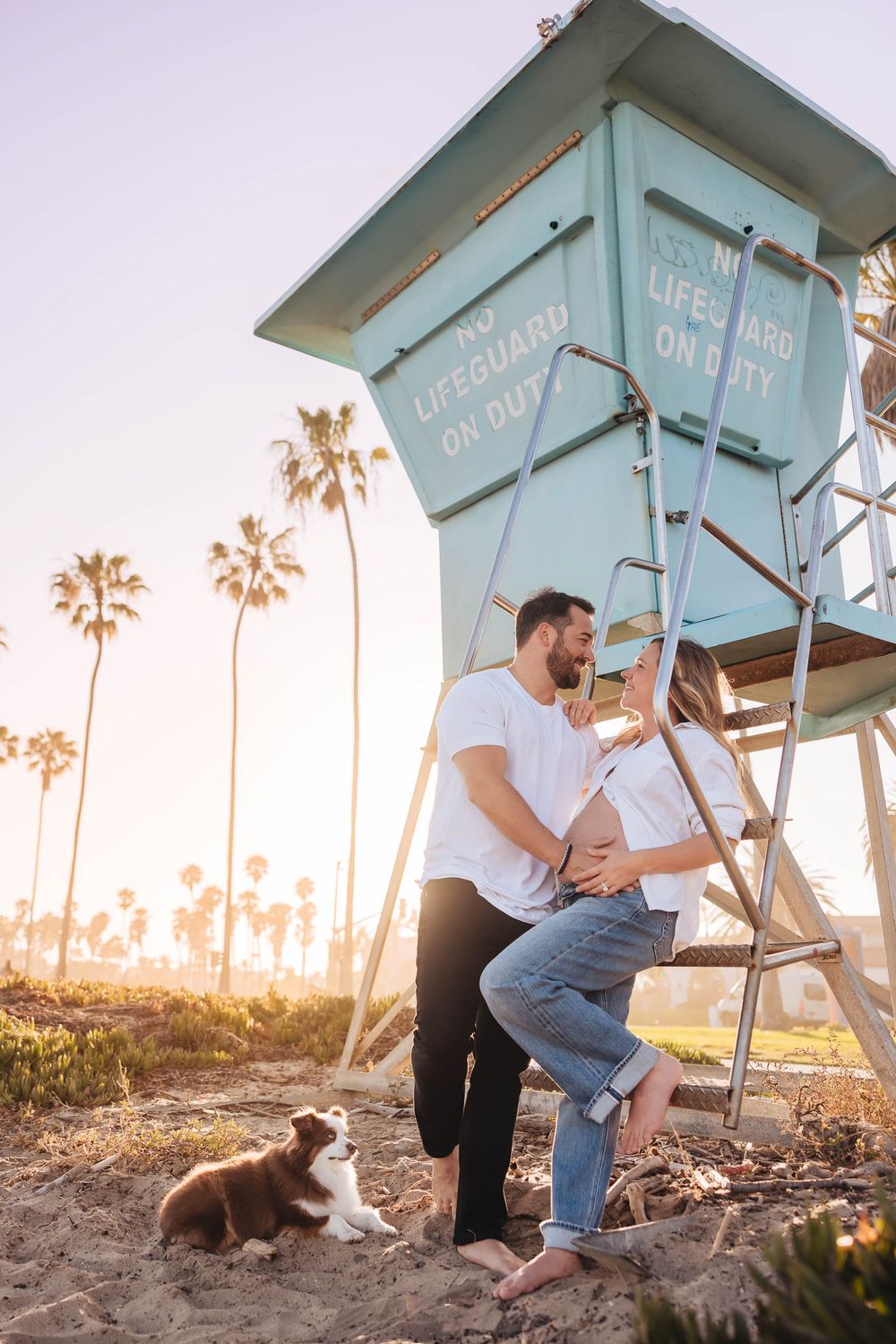 Pregnant couple embracing near teal lifeguard tower on sunny beach with palm trees and brown dog nearby.