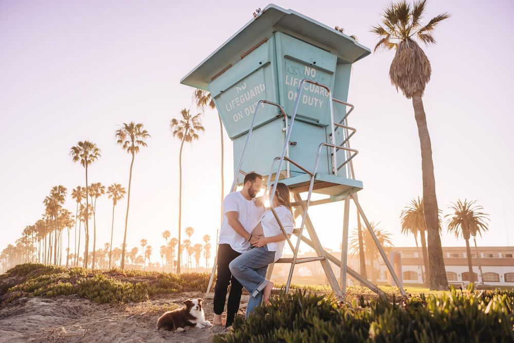 Couple sitting together by teal lifeguard tower on sandy beach surrounded by tall palm trees at sunset. Maternity photos Santa Barbara