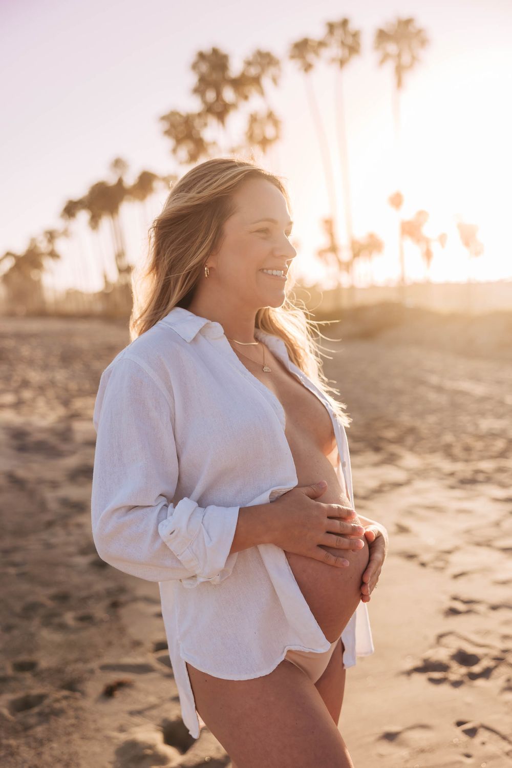 Smiling pregnant woman in white shirt holding belly on sunny beach at golden hour. Client Tips