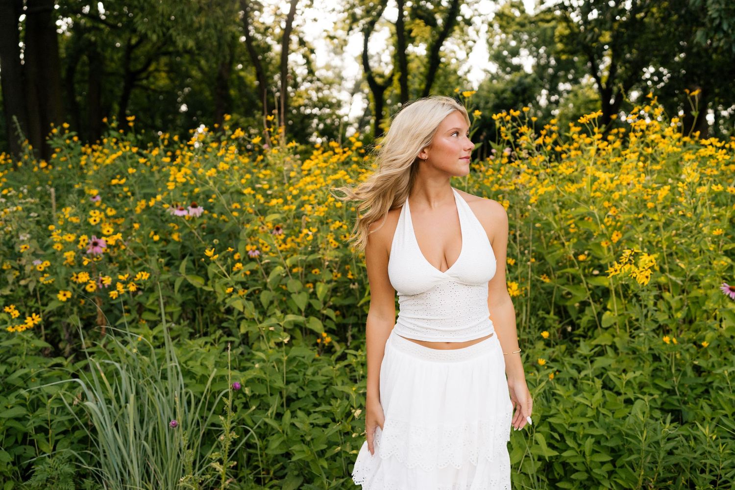 Smiling blonde woman in white halter top and tiered skirt standing in a sunlit meadow with trees behind her.