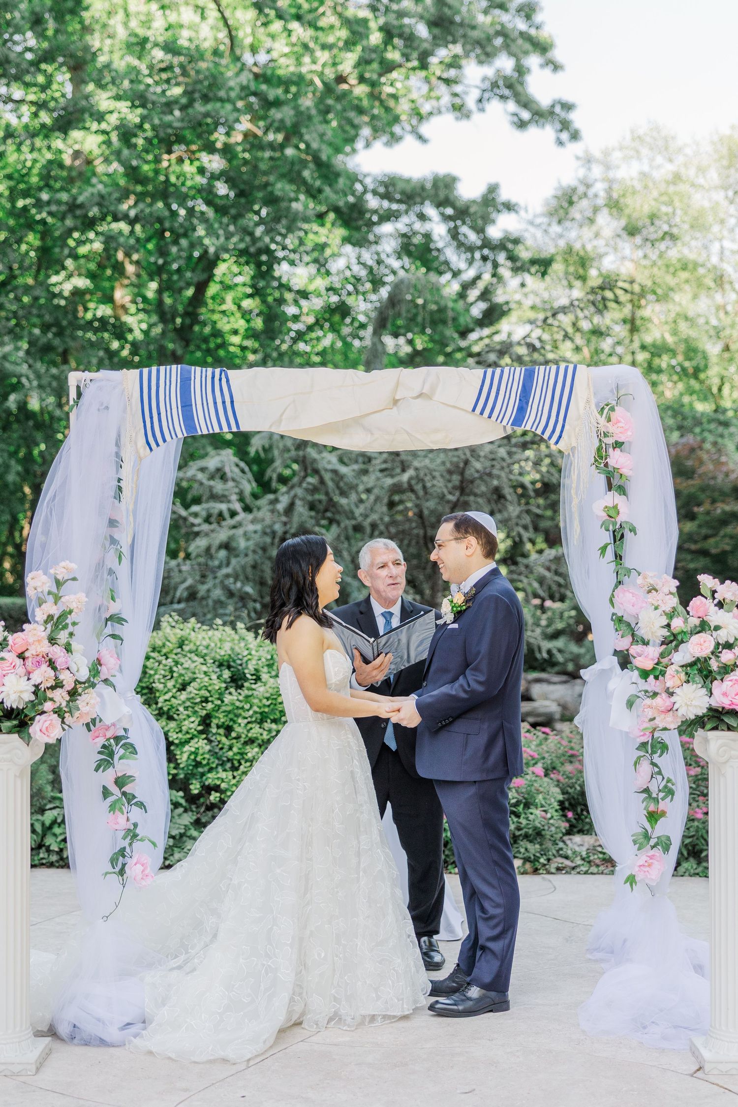 Couple exchanges vows under a floral chuppah with pink roses and draped fabric at an outdoor Jewish wedding ceremony.