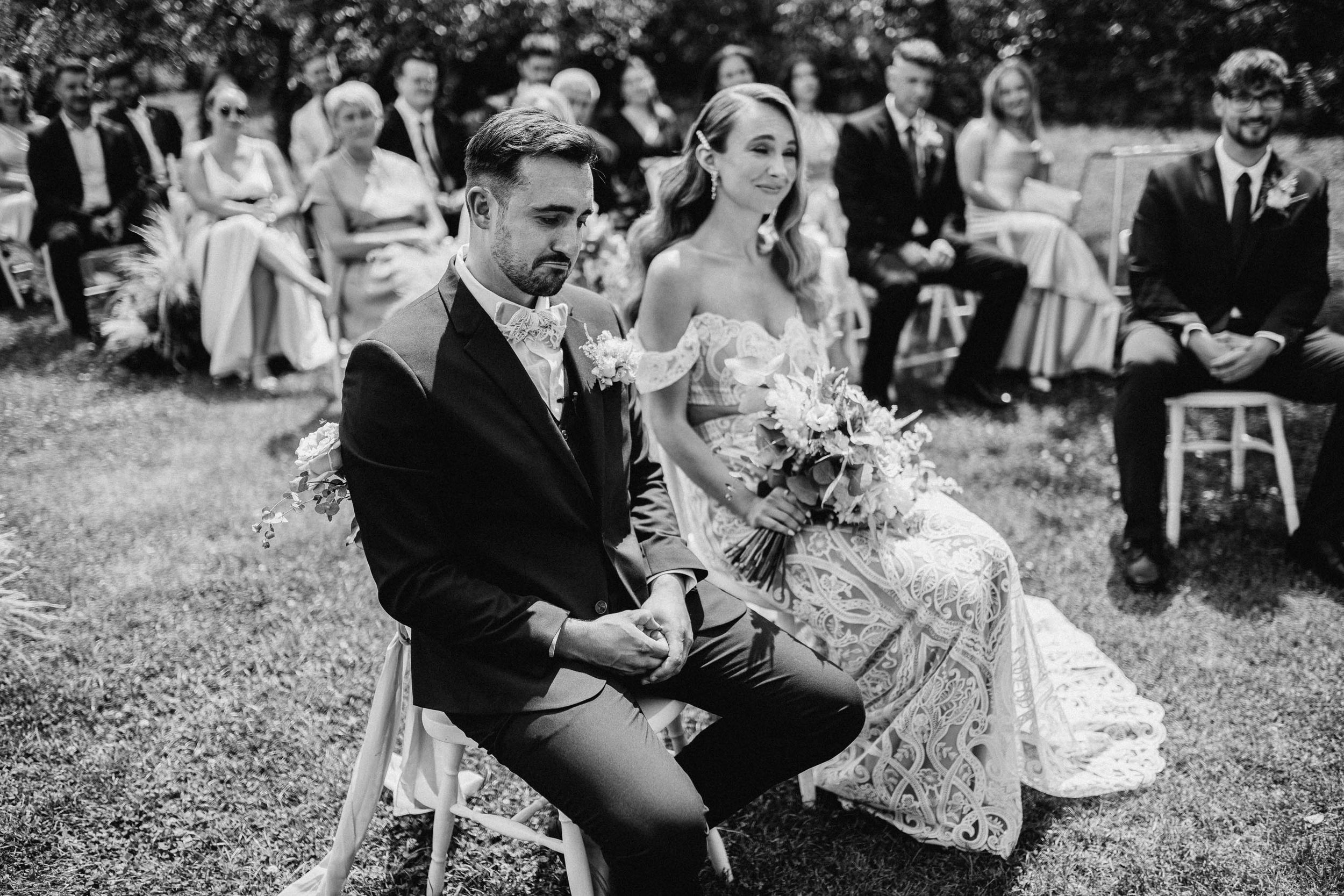 Černobílá fotografie ženicha s pejskem mezi hosty při venkovním obřadu – Statek Vitice, Pavlína a Vojta. Svatební fotograf Jiří Tulach.